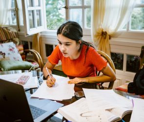 hispanic latina college student works on assignment at home. She is writing something on paper. A laptop is on her desk.