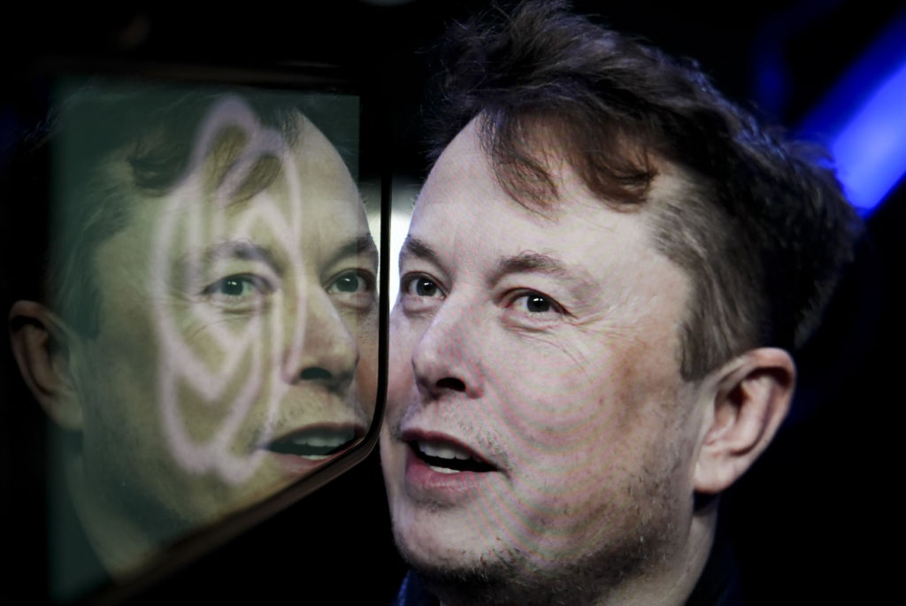 In this photo illustration, the logo of 'OpenAI' is displayed on a mobile phone screen in front of a computer screen displaying the photograph of Elon Musk.