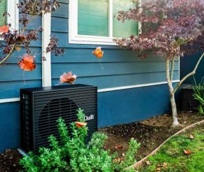 A dark grey heat pump sits outside a blue home.