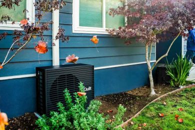 A dark grey heat pump sits outside a blue home.