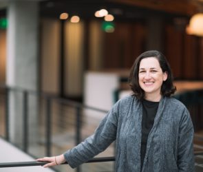 A dark-haired woman poses by a balcony in a gray cardigan sweater.