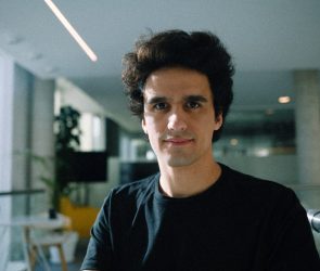 A man wearing a large black t-shirt stands in a sunlit office.