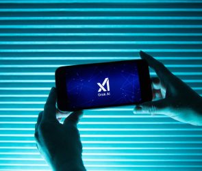 A person holds a telephone displaying the logo of Elon Musk s artificial intelligence grok in front of a background lit by a blue light.