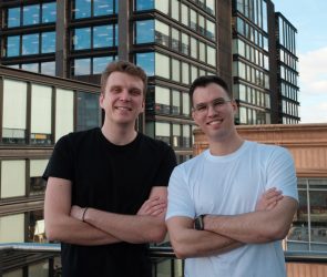 Two men stand on a rooftop, crossing their arms