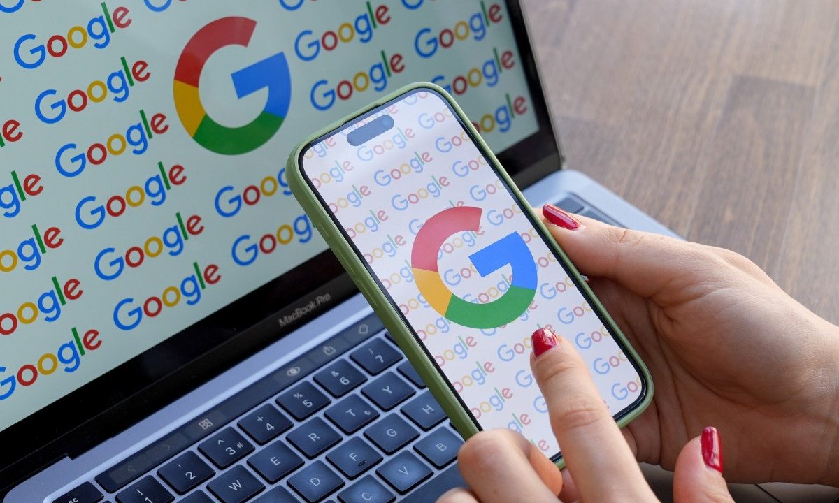 A woman looks at a mobile phone displaying the logo of Google in front of a laptop screen displaying the logo of Google.