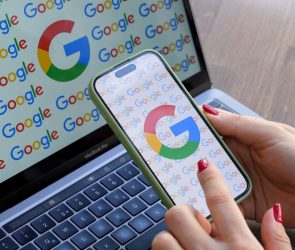 A woman looks at a mobile phone displaying the logo of Google in front of a laptop screen displaying the logo of Google.